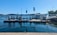 A waterfront scene featuring a fuel dock on a clear day. The dock is equipped with several fuel pumps and is positioned in a harbor area. A small building with colorful flags on the roof is visible, and beyond the dock, calm water extends towards a distant treeline. Boats can be seen in the background, along with clear blue skies overhead.