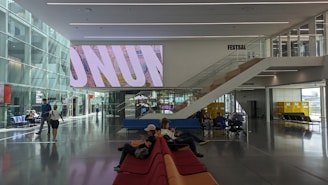 A modern indoor space with high ceilings and large glass windows, featuring multiple people sitting and walking. There are colorful couches and a large digital screen displaying text. An open area with stairs leading up to another level and some yellow lockers can be seen in the background.