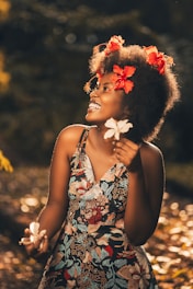 A woman is smiling joyfully while standing in a sunlit area. She wears a floral dress with intricate patterns and vibrant colors. Red flowers adorn her curly hair, and she holds white flowers in her hands. The background is softly blurred, suggesting an outdoor setting with dappled sunlight and natural surroundings.