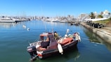 A small motorboat is docked in a calm marina with clear blue water. Several yachts are lined up across the water, with a cityscape of mid-rise buildings in the background. The sky is clear and sunny, and a few palm trees and green spaces are visible by the waterfront.