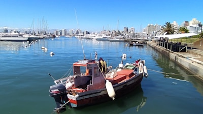A small motorboat is docked in a calm marina with clear blue water. Several yachts are lined up across the water, with a cityscape of mid-rise buildings in the background. The sky is clear and sunny, and a few palm trees and green spaces are visible by the waterfront.