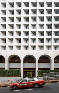 A modern building facade featuring a geometric pattern with recessed windows creating a grid-like appearance. The lower part of the building has large arches and a minimalistic design, accompanied by neatly trimmed greenery. A red taxi is captured in motion in the foreground on a city street.
