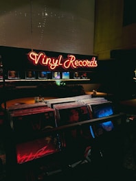 A dimly lit record store with a glowing neon sign displaying 'Vinyl Records' in red. The foreground shows a collection of vinyl records, with some album covers partially visible under low light. The atmosphere is cozy and nostalgic, typical of vintage or retro-themed shops.