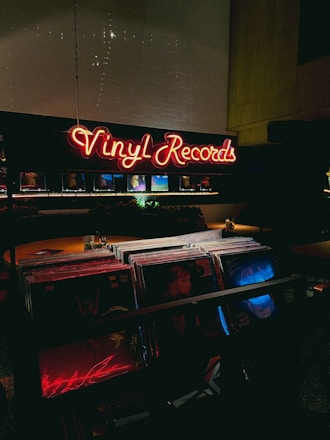 A dimly lit record store with a glowing neon sign displaying 'Vinyl Records' in red. The foreground shows a collection of vinyl records, with some album covers partially visible under low light. The atmosphere is cozy and nostalgic, typical of vintage or retro-themed shops.