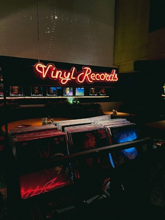 A dimly lit record store with a glowing neon sign displaying 'Vinyl Records' in red. The foreground shows a collection of vinyl records, with some album covers partially visible under low light. The atmosphere is cozy and nostalgic, typical of vintage or retro-themed shops.