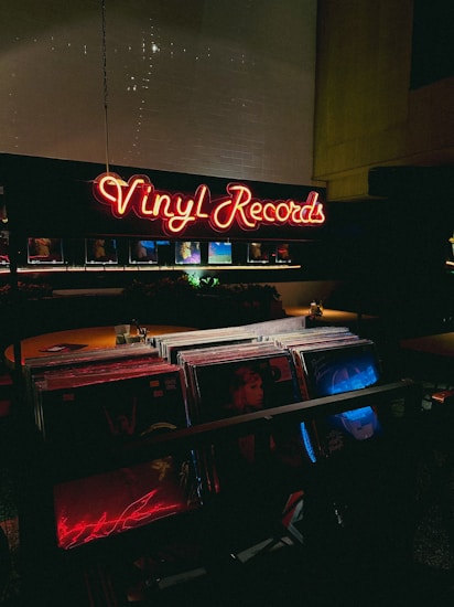 A dimly lit record store with a glowing neon sign displaying 'Vinyl Records' in red. The foreground shows a collection of vinyl records, with some album covers partially visible under low light. The atmosphere is cozy and nostalgic, typical of vintage or retro-themed shops.