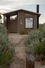 A small, outdoor public restroom facility made of concrete, situated along a dirt pathway surrounded by dense green shrubbery. The structure features a roof and a notice board displaying various information. Nearby signs indicate accessibility for people with disabilities and the presence of a toilet. A long black vent pipe extends above the roof on the right side.