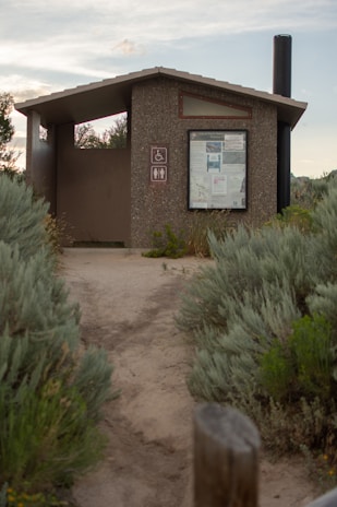 A small, outdoor public restroom facility made of concrete, situated along a dirt pathway surrounded by dense green shrubbery. The structure features a roof and a notice board displaying various information. Nearby signs indicate accessibility for people with disabilities and the presence of a toilet. A long black vent pipe extends above the roof on the right side.