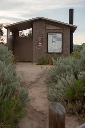 A small, outdoor public restroom facility made of concrete, situated along a dirt pathway surrounded by dense green shrubbery. The structure features a roof and a notice board displaying various information. Nearby signs indicate accessibility for people with disabilities and the presence of a toilet. A long black vent pipe extends above the roof on the right side.