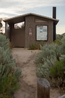 A small, outdoor public restroom facility made of concrete, situated along a dirt pathway surrounded by dense green shrubbery. The structure features a roof and a notice board displaying various information. Nearby signs indicate accessibility for people with disabilities and the presence of a toilet. A long black vent pipe extends above the roof on the right side.