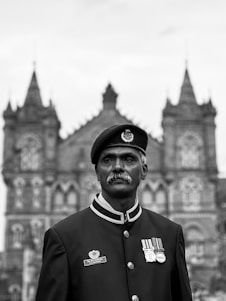 A uniformed figure stands in front of a historic building with intricate architectural details, including spires and arched windows. The figure wears a beret adorned with an insignia and has several medals on the chest.