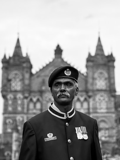 A uniformed figure stands in front of a historic building with intricate architectural details, including spires and arched windows. The figure wears a beret adorned with an insignia and has several medals on the chest.