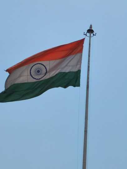A flag is flying on a tall pole against a clear blue sky. The flag features horizontal stripes of orange, white, and green with a blue wheel symbol in the center of the white stripe.