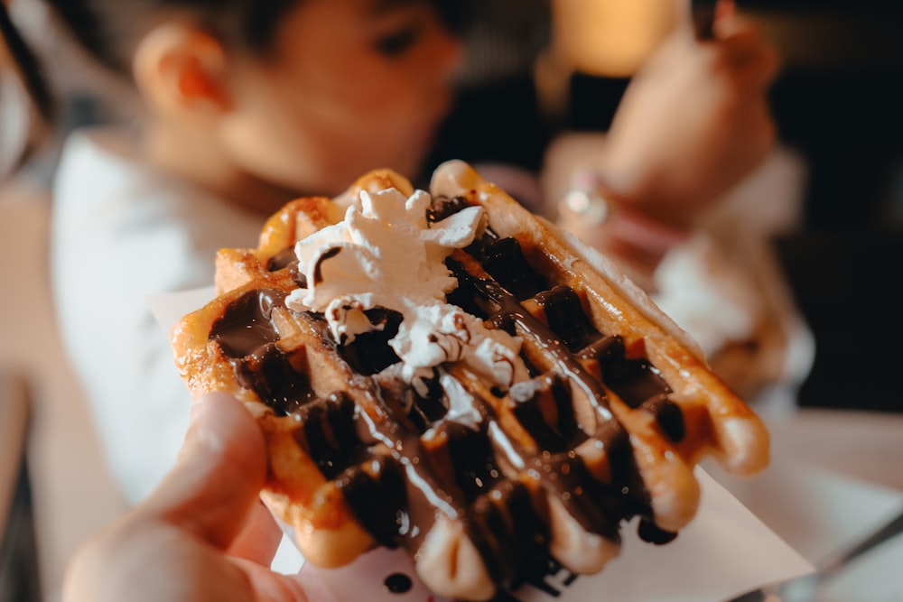a person holding a waffle covered in chocolate and whipped cream