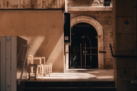 A narrow architectural passageway with an arched doorway made of stone and dark wood details leads into a darker interior. There are shadows and light patterns on the ground from the afternoon sun. On the left side, there are white chairs and a table against a beige wall. The surroundings consist of stone walls and a mix of modern and old elements.
