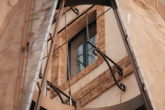 A stone-framed window with rustic metal railings, surrounded by beige and brown tones. The scene includes metallic structures and fabric elements partially visible around the window frame.