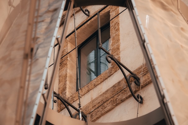 A stone-framed window with rustic metal railings, surrounded by beige and brown tones. The scene includes metallic structures and fabric elements partially visible around the window frame.