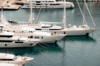 Several yachts are docked at a marina with clear, calm blue water. There are multiple white luxury boats lined up next to each other, and a few people can be seen on the decks. In the background, there is a larger yacht farther away, with small orange fenders hanging on its side. There are also a few smaller boats and a part of the marina dock visible.
