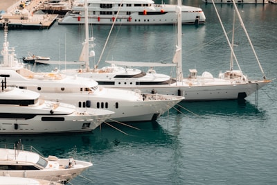 Several yachts are docked at a marina with clear, calm blue water. There are multiple white luxury boats lined up next to each other, and a few people can be seen on the decks. In the background, there is a larger yacht farther away, with small orange fenders hanging on its side. There are also a few smaller boats and a part of the marina dock visible.