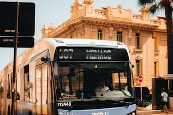 A modern street bus traveling through an urban area with historical architecture. The bus is white with blue accents and has digital signage displaying information. The background shows a classic European-style building with intricate details and a palm tree to the side.