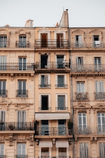 A multi-story residential building with ornate balconies and a neutral color palette. The architecture features intricate wrought iron railings and classical design elements. Several windows and balconies are visible, with some open or partially closed. The exterior walls are painted in light beige and tan tones.