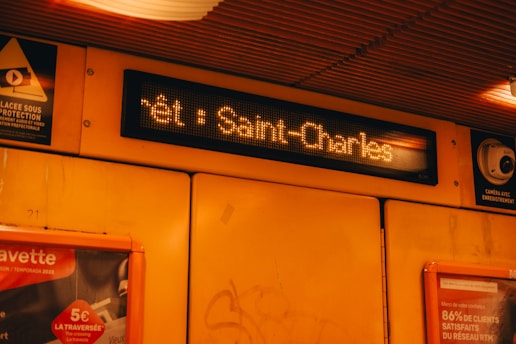 An interior view of a subway or train station with a digital display showing 'Saint-Charles'. The walls are yellow with various posters and advertisements. A surveillance camera is mounted to the right.