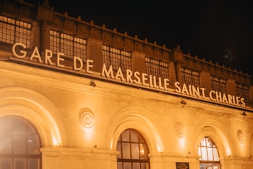 The architectural facade of a train station is illuminated at night. Large, bold letters spell out 'Gare de Marseille Saint Charles' on top of the building, while warm, yellow lights highlight its decorative stone carvings and arched windows.