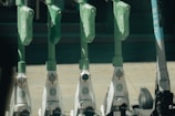 A row of electric scooters parked in a line, each equipped with a green handlebar and steering column. The scooters are white at the bottom with Uber branding, except for one on the far right with Bird branding. The ground appears to be made of paved stone, and there is a dark, blurry background.