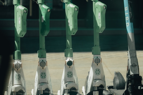 A row of electric scooters parked in a line, each equipped with a green handlebar and steering column. The scooters are white at the bottom with Uber branding, except for one on the far right with Bird branding. The ground appears to be made of paved stone, and there is a dark, blurry background.