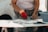 A person is cleaning fish on a white table using a knife. They are wearing a floral-patterned shirt and a red glove on one hand. Next to them is a gray plastic container and some other utensils on the table.