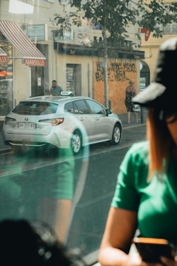 A street scene featuring a parked silver taxi with a visible license plate. The background includes a building with a red and white striped awning labeled 'MARSEILLE', graffiti on boarded windows, and a couple of pedestrians walking. A person in the foreground inside a vehicle is partially visible, wearing a green shirt and black cap, holding a phone.