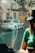A street scene featuring a parked silver taxi with a visible license plate. The background includes a building with a red and white striped awning labeled 'MARSEILLE', graffiti on boarded windows, and a couple of pedestrians walking. A person in the foreground inside a vehicle is partially visible, wearing a green shirt and black cap, holding a phone.