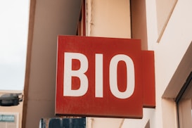 A red sign with the white letters 'BIO' mounted on a wall. The sign appears to be part of a storefront or business facade, surrounded by beige-colored walls.