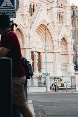 A large, ornate building with intricate stone carvings and arched doorways dominates the background. Two people are walking near a construction sign that mentions renovation. A person in a red shirt is seen in the foreground near a street sign and traffic pole. The scene appears to be urban, with visible overhead wires and a street.