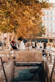 A lively urban scene with people walking near a tree-lined street. A sign for the Charles de Gaulle metro station is visible at the forefront, with autumn foliage overhead.