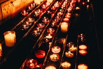 Numerous lit candles in small glass holders are arranged in rows, creating a warm and serene atmosphere. The candles emit a soft glow, casting reflections and shadows against a dark background.