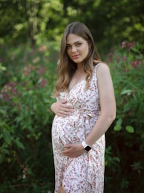 A pregnant woman stands in a lush green garden, wearing a light floral dress. Her hands are gently placed on her belly. She has long brown hair and is looking at the camera, exuding a serene and peaceful expression.