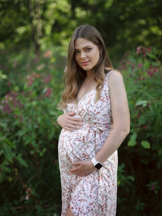 A pregnant woman stands in a lush green garden, wearing a light floral dress. Her hands are gently placed on her belly. She has long brown hair and is looking at the camera, exuding a serene and peaceful expression.