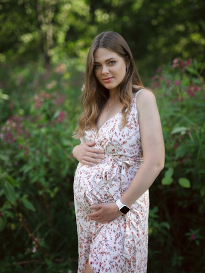 A pregnant woman stands in a lush green garden, wearing a light floral dress. Her hands are gently placed on her belly. She has long brown hair and is looking at the camera, exuding a serene and peaceful expression.