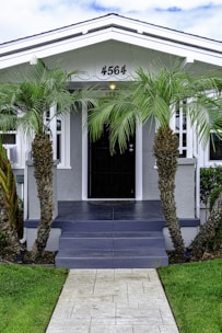 A charming, single-story house with a gabled roof and a front porch adorned by multiple palm trees. The front pathway is neatly paved, leading to a dark-colored door framed by light gray walls. White-framed windows are symmetrically placed on either side of the doorway.