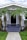 A charming, single-story house with a gabled roof and a front porch adorned by multiple palm trees. The front pathway is neatly paved, leading to a dark-colored door framed by light gray walls. White-framed windows are symmetrically placed on either side of the doorway.