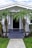 A charming, single-story house with a gabled roof and a front porch adorned by multiple palm trees. The front pathway is neatly paved, leading to a dark-colored door framed by light gray walls. White-framed windows are symmetrically placed on either side of the doorway.