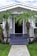 A charming, single-story house with a gabled roof and a front porch adorned by multiple palm trees. The front pathway is neatly paved, leading to a dark-colored door framed by light gray walls. White-framed windows are symmetrically placed on either side of the doorway.