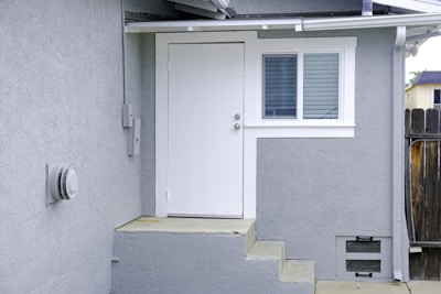 A small exterior entryway features a white door with a metal doorknob, adjacent to a double-paned window with white trim. The walls are painted light gray, and there is a short set of concrete steps leading up to the door. A wooden fence is visible on the right, and a utility meter is mounted on the left wall.