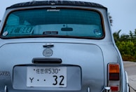 Rear view of a classic Mini Cooper with raindrops on its surface. The car has multiple license plates, one displaying Japanese characters. A sticker indicating 'Rover Japan' is visible on the rear windshield. The background shows some greenery.