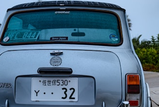 Rear view of a classic Mini Cooper with raindrops on its surface. The car has multiple license plates, one displaying Japanese characters. A sticker indicating 'Rover Japan' is visible on the rear windshield. The background shows some greenery.