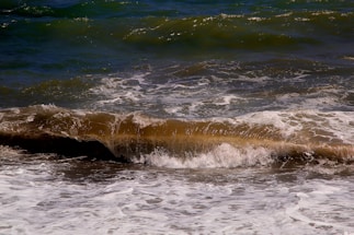 A dynamic ocean scene features waves crashing against each other with foamy white edges. The water displays a mixture of deep blue and brown hues, indicating possible sediments or stirred sand.