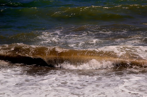A dynamic ocean scene features waves crashing against each other with foamy white edges. The water displays a mixture of deep blue and brown hues, indicating possible sediments or stirred sand.