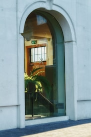 Arched window with a glass pane. Inside, a staircase leading downwards is visible, along with a potted fern plant. Light from a large rectangular window illuminates the room. An exit sign is mounted above a door in the background.