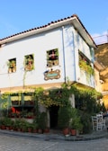 A charming café located on a cobblestone street, with a rustic two-story building adorned with vibrant green plants and flowers cascading from window boxes. The café features potted plants outside, adding to the cozy and inviting atmosphere. A sign hangs above the entrance, and a few chairs and tables are set up outside for patrons to enjoy.